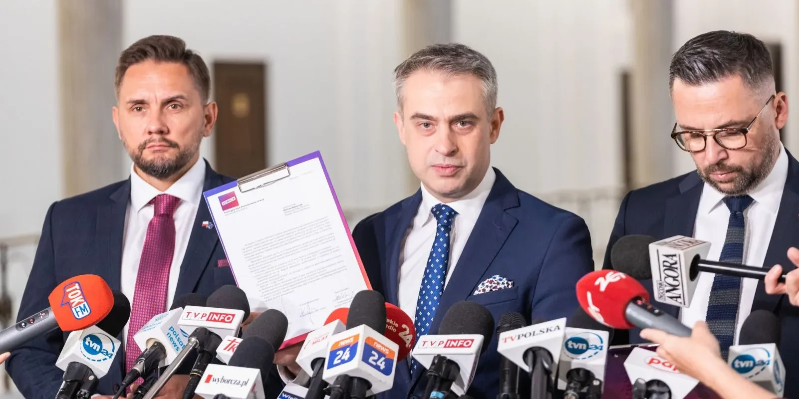 Trzech mężczyzn w garniturach podczas konferencji prasowej, stojących za licznymi mikrofonami; jeden z nich trzyma dokument w fioletowej teczce.