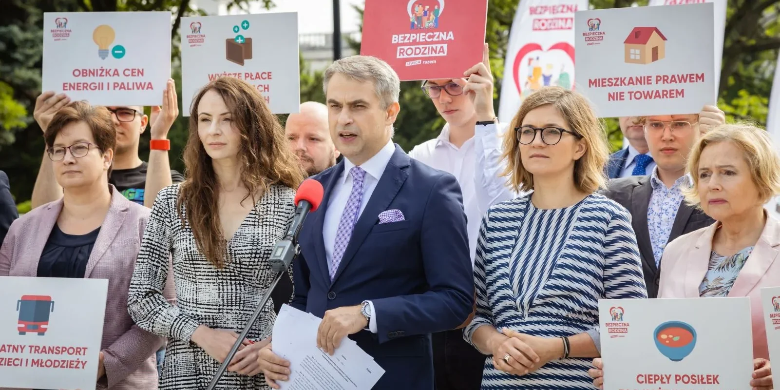 Na zdjęciu widoczny jest mężczyzna mówiący do mikrofonu, stojący wśród grupy osób trzymających transparenty dotyczące różnych postulatów społecznych.