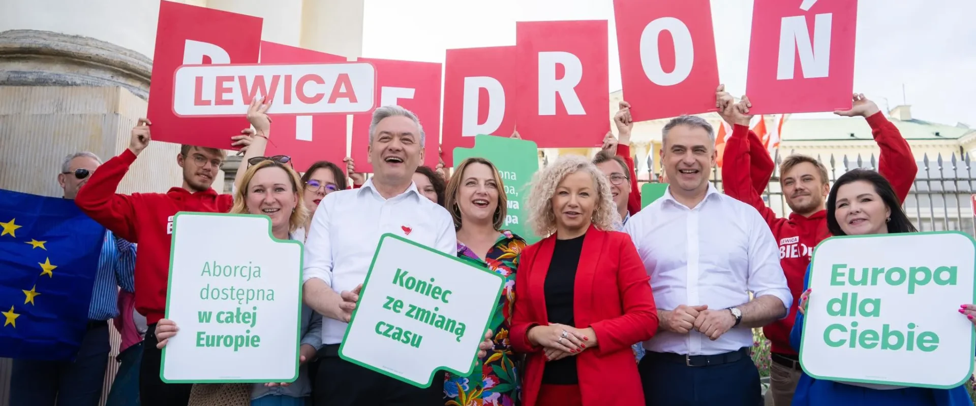 Grupa zwolenników Lewicy podczas demonstracji trzyma plakaty z hasłami „Europa dla Ciebie”, „Koniec ze zmianą czasu” i „Aborcja dostępna w całej Europie” na tle kolumn i ogrodzenia.