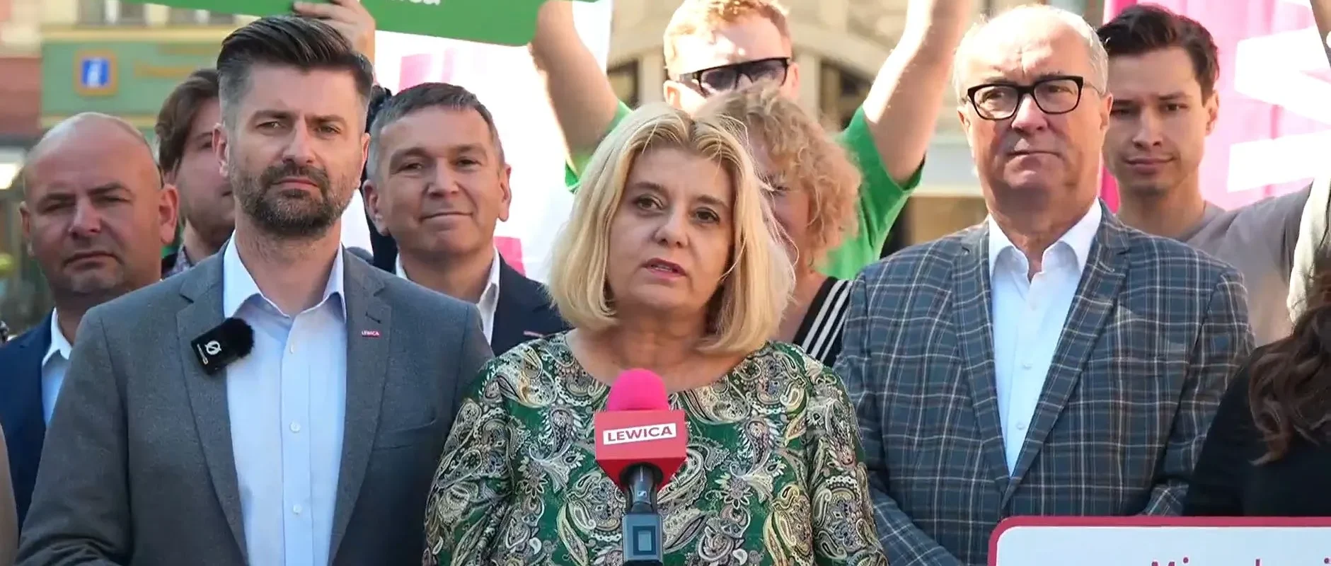 Kobieta o blond włosach przemawia do mikrofonu z logo Lewicy podczas wiecu, otoczona grupą zwolenników i plakatami w tle.