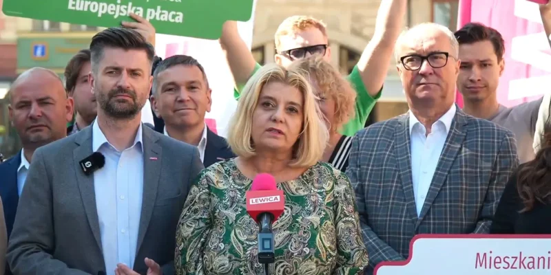 Kobieta o blond włosach przemawia do mikrofonu z logo Lewicy podczas wiecu, otoczona grupą zwolenników i plakatami w tle.