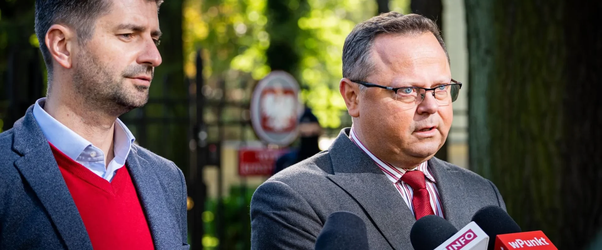 Dwóch mężczyzn w garniturach podczas konferencji prasowej w parku, stojących przy mikrofonach z logotypami TVN24, TVP Info i WPunkt.