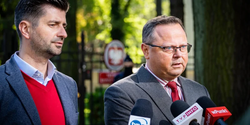 Dwóch mężczyzn w garniturach podczas konferencji prasowej w parku, stojących przy mikrofonach z logotypami TVN24, TVP Info i WPunkt.