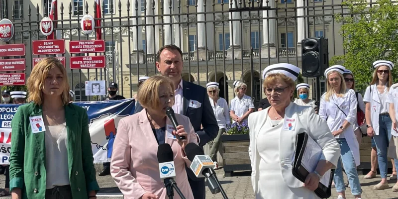 Grupa pielęgniarek i położnych w białych fartuchach i czapkach pielęgniarskich stoi przed kolumnowym budynkiem z metalową balustradą i wygłasza przemówienie podczas ostrzegawczego strajku w Warszawie.