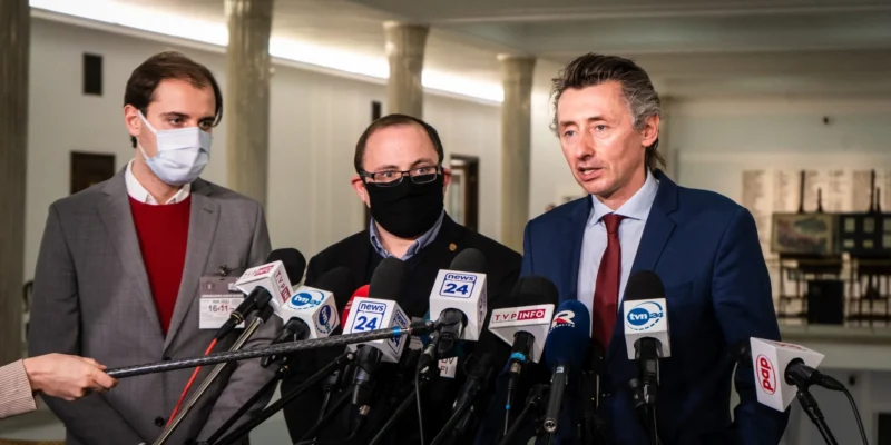 Na zdjęciu trzech mężczyzn stoi przed mikrofonami podczas konferencji prasowej w budynku, prawdopodobnie rządowym.