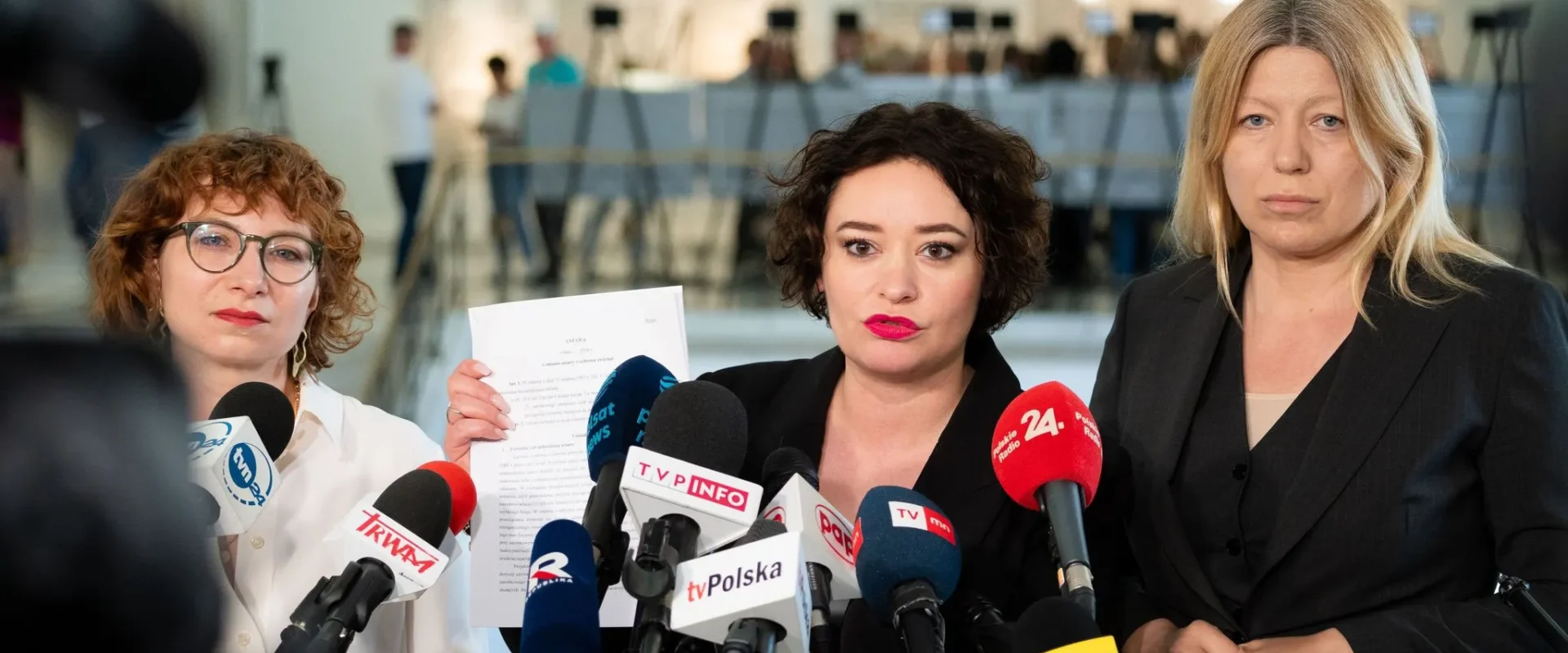 Trzy kobiety stoją przy mikrofonach na konferencji prasowej; jedna z nich trzyma dokument, a w tle widać salę z ludźmi.