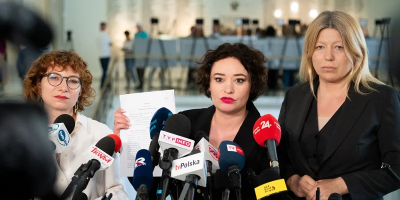 Trzy kobiety stoją przy mikrofonach na konferencji prasowej; jedna z nich trzyma dokument, a w tle widać salę z ludźmi.