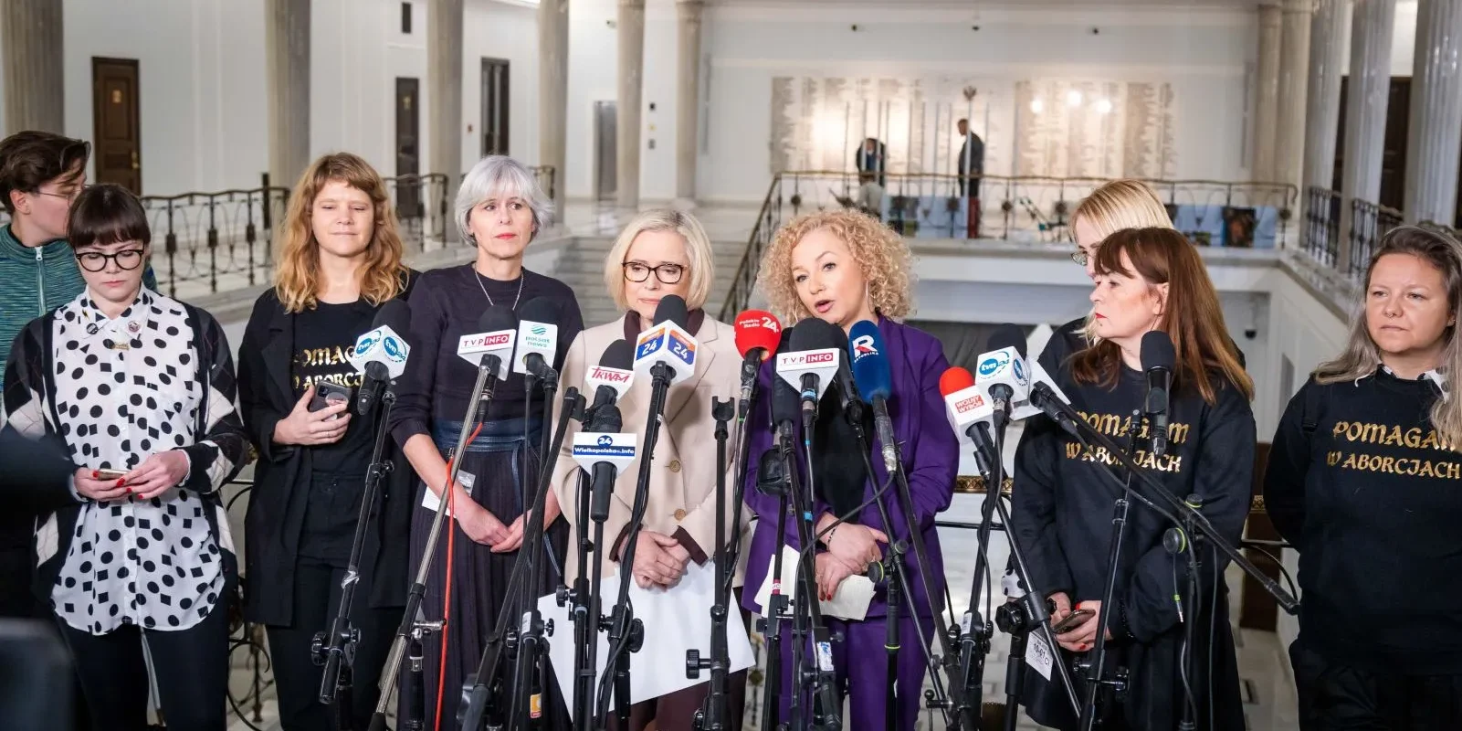 Na zdjęciu widocznych jest osiem kobiet stojących przed mikrofonami, z których część ma na sobie koszulki z napisem "Pomagam w aborcjach", podczas konferencji prasowej w eleganckim wnętrzu.