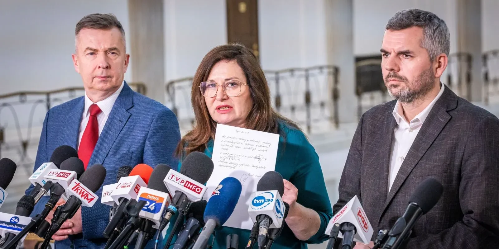 Trójka polityków podczas konferencji prasowej, otoczona licznymi mikrofonami, kobieta w środku trzyma kartkę i wygłasza oświadczenie w sprawie odblokowania ustawy wiatrakowej.