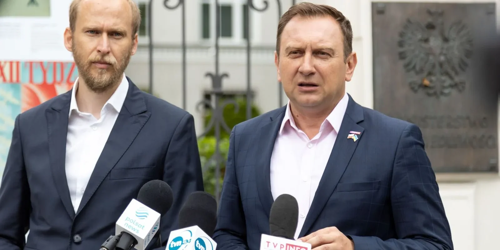 Dwóch mężczyzn w garniturach stoi przy mikrofonach podczas konferencji prasowej, z widocznymi logotypami mediów.