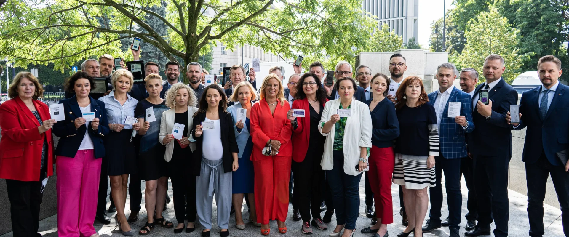 Grupa parlamentarzystów Lewicy na zewnątrz pod drzewem, wszyscy trzymają kartę lub dokument potwierdzający szczepienie przeciw COVID-19.