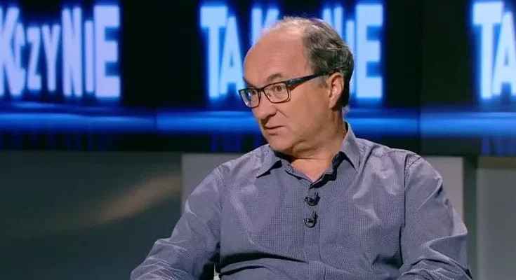 Mężczyzna w średnim wieku w koszuli i okularach siedzi w studiu telewizyjnym z przypiętym mikrofonem i rozmawia.