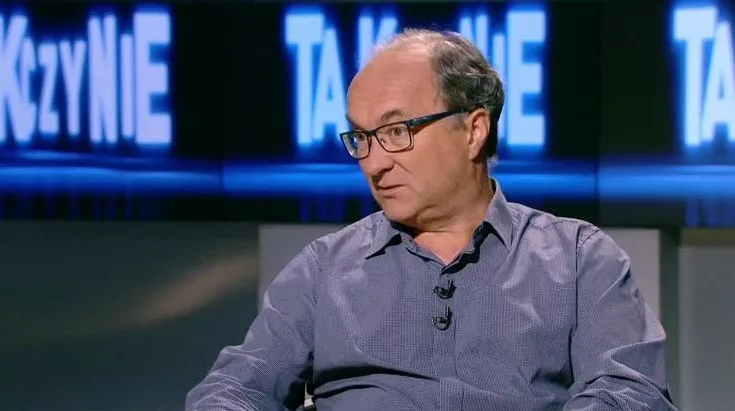 Mężczyzna w średnim wieku w koszuli i okularach siedzi w studiu telewizyjnym z przypiętym mikrofonem i rozmawia.