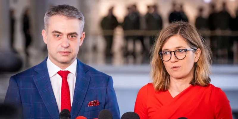 Dwoje ludzi w formalnym ubraniu stoi przed mikrofonami, przemawiając na konferencji prasowej.