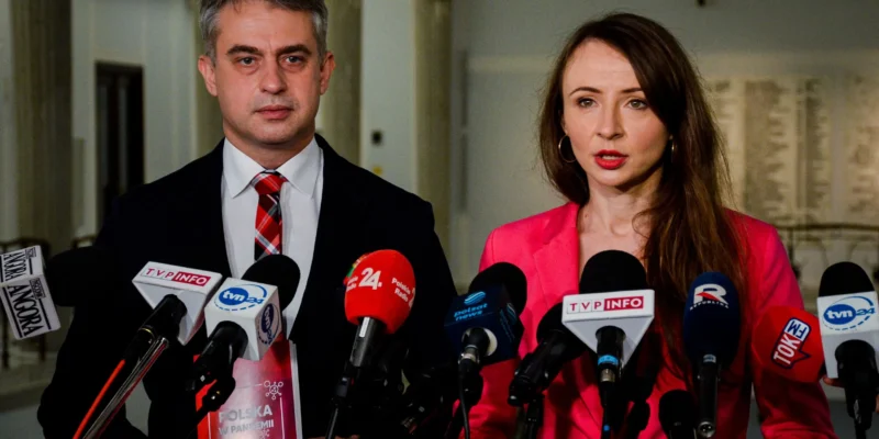 Dwóch polityków podczas konferencji prasowej, otoczonych licznymi mikrofonami mediów, omawia temat czwartej fali koronawirusa.