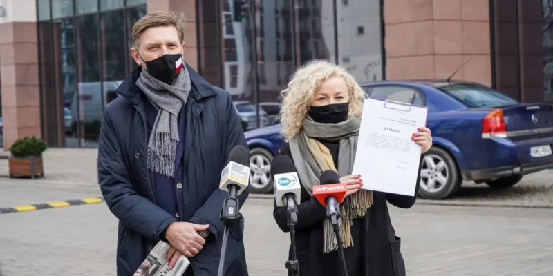 Dwoje osób w maseczkach stoi przy mikrofonach i trzyma dokument podczas konferencji prasowej na zewnątrz budynku.