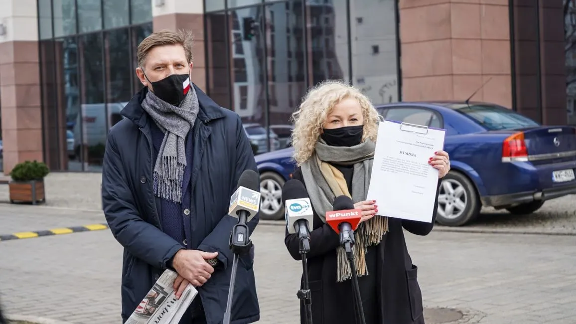 Dwoje osób w maseczkach stoi przy mikrofonach i trzyma dokument podczas konferencji prasowej na zewnątrz budynku.