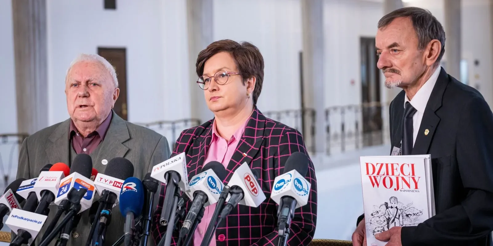 Trzy osoby stojące podczas konferencji prasowej w hali z kolumnami, wokół licznych mikrofonów; po lewej starszy mężczyzna, w środku kobieta w okularach i różowej koszuli w kratkę, a po prawej mężczyzna trzymający publikację z tytułem „Dzieci Wojny”.