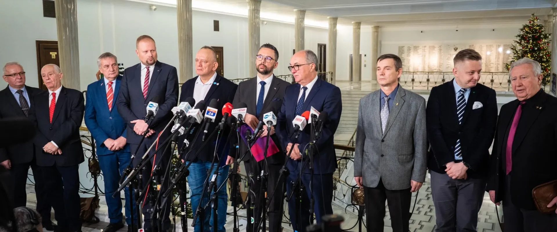 Na zdjęciu znajduje się grupa mężczyzn w garniturach, stojących w półokręgu przed mikrofonami w eleganckim wnętrzu, dekorowanym świątecznym drzewkiem.