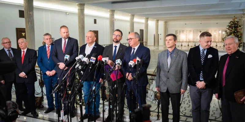 Na zdjęciu znajduje się grupa mężczyzn w garniturach, stojących w półokręgu przed mikrofonami w eleganckim wnętrzu, dekorowanym świątecznym drzewkiem.