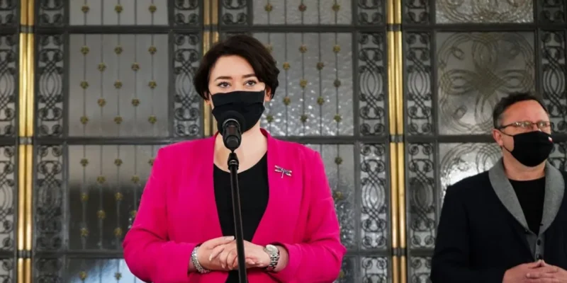 Kobieta w różowej marynarce i maseczce stoi przy mikrofonie, a obok niej mężczyzna w czerni i maseczce, na tle ozdobnej metalowej bramy.