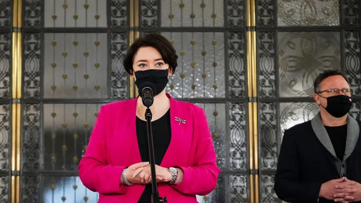 Kobieta w różowej marynarce i maseczce stoi przy mikrofonie, a obok niej mężczyzna w czerni i maseczce, na tle ozdobnej metalowej bramy.