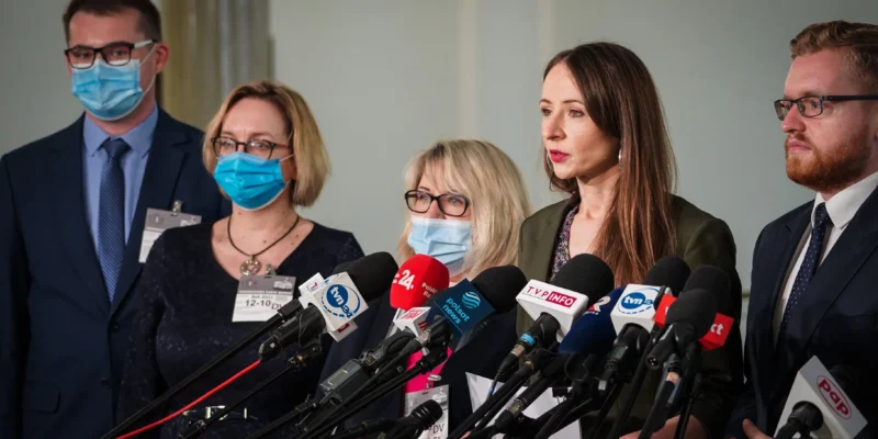 Na zdjęciu przedstawiono grupę czterech osób w maseczkach ochronnych, stojących przy mikrofonach podczas konferencji prasowej.