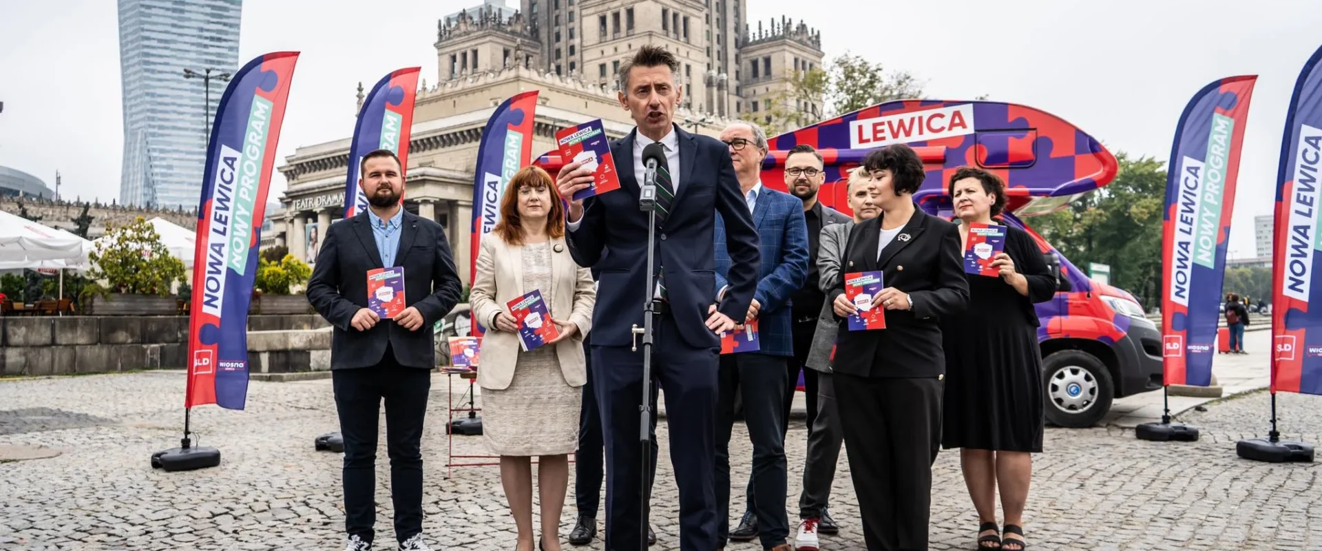 Grupa polityków Nowej Lewicy stoi na placu podczas konferencji prasowej, trzyma ulotki i stoi przy mikrofonie, z w tle widoczne flagi i banery Nowa Lewica oraz czerwony pojazd reklamowy.