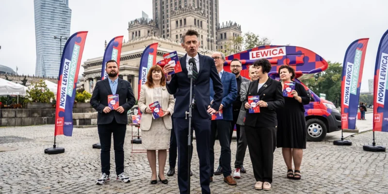 Grupa polityków Nowej Lewicy stoi na placu podczas konferencji prasowej, trzyma ulotki i stoi przy mikrofonie, z w tle widoczne flagi i banery Nowa Lewica oraz czerwony pojazd reklamowy.