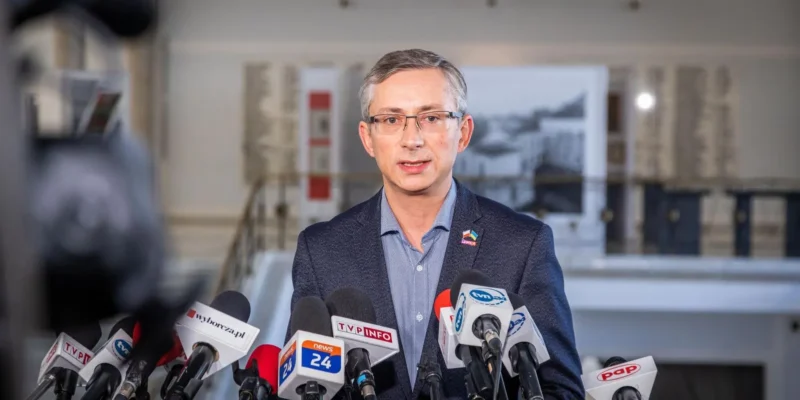 Na zdjęciu przedstawiony jest mężczyzna w okularach, przemawiający do dziennikarzy przed mikrofoniami w sali konferencyjnej.