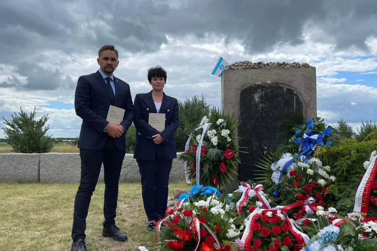 Na zdjęciu widoczni są dwie osoby stojące obok pomnika, otoczonego kwiatami i flagą, w scenerii z pochmurnym niebem.