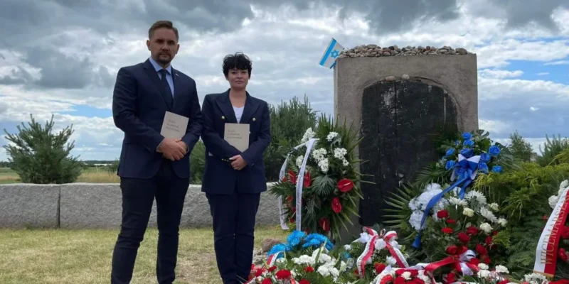 Na zdjęciu widoczni są dwie osoby stojące obok pomnika, otoczonego kwiatami i flagą, w scenerii z pochmurnym niebem.