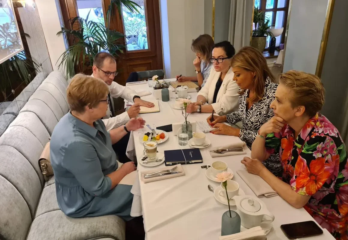 Grupa osób siedzi przy eleganckim stole w restauracji, rozmawiając i notując, otoczona roślinnością oraz subtelnym wystrojem wnętrza.