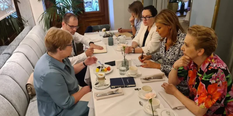 Grupa osób siedzi przy eleganckim stole w restauracji, rozmawiając i notując, otoczona roślinnością oraz subtelnym wystrojem wnętrza.