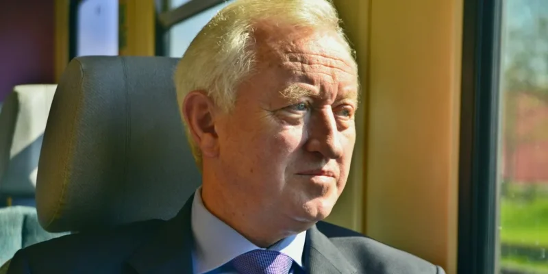 Na zdjęciu widoczny jest mężczyzna w garniturze, siedzący przy oknie, z zamyślonym wyrazem twarzy.