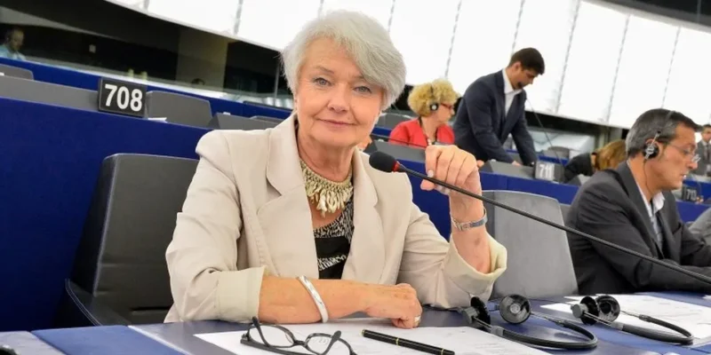 Kobieta w jasnej marynarce siedzi przy mikrofonie w sali obrad, patrząc przed siebie, a wokół niej inni parlamentarzyści z zestawami słuchawkowymi.