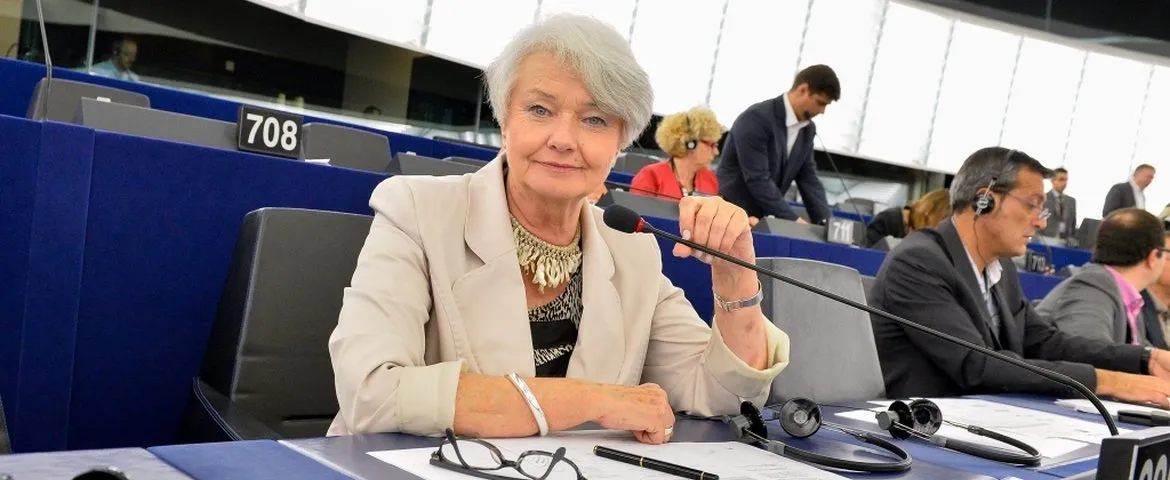 Kobieta w jasnej marynarce siedzi przy mikrofonie w sali obrad, patrząc przed siebie, a wokół niej inni parlamentarzyści z zestawami słuchawkowymi.