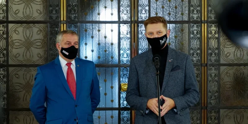 Dwóch mężczyzn w garniturach i maseczkach stoi przed ozdobnymi drzwiami, a jeden z nich przemawia do mikrofonu.