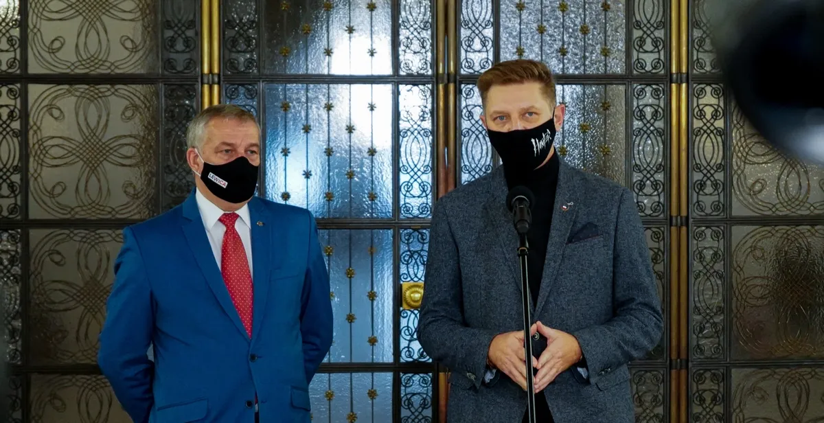 Dwóch mężczyzn w garniturach i maseczkach stoi przed ozdobnymi drzwiami, a jeden z nich przemawia do mikrofonu.