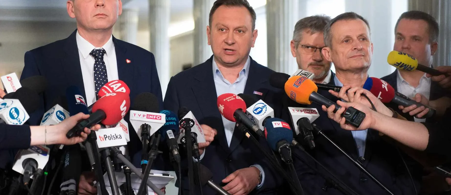 Grupa mężczyzn w garniturach udziela konferencji prasowej, stojąc wśród licznych mikrofonów skierowanych do reporterów.