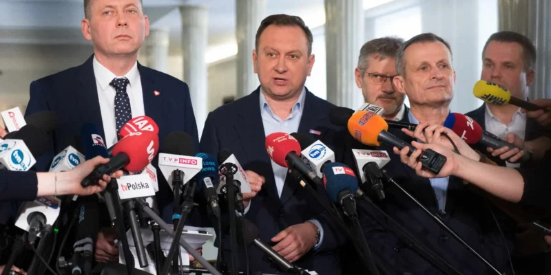 Grupa mężczyzn w garniturach udziela konferencji prasowej, stojąc wśród licznych mikrofonów skierowanych do reporterów.