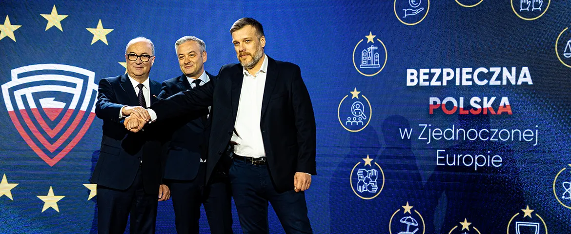 Na zdjęciu trzech mężczyzn stoi razem na scenie, uśmiechając się i wymieniając uściski dłoni, w tle widoczny jest baner z napisem "Bezpieczna Polska w Zjednoczonej Europie".