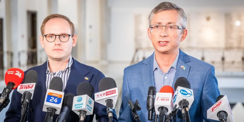 Dwóch mężczyzn w garniturach stoi przy mikrofonach podczas konferencji prasowej.