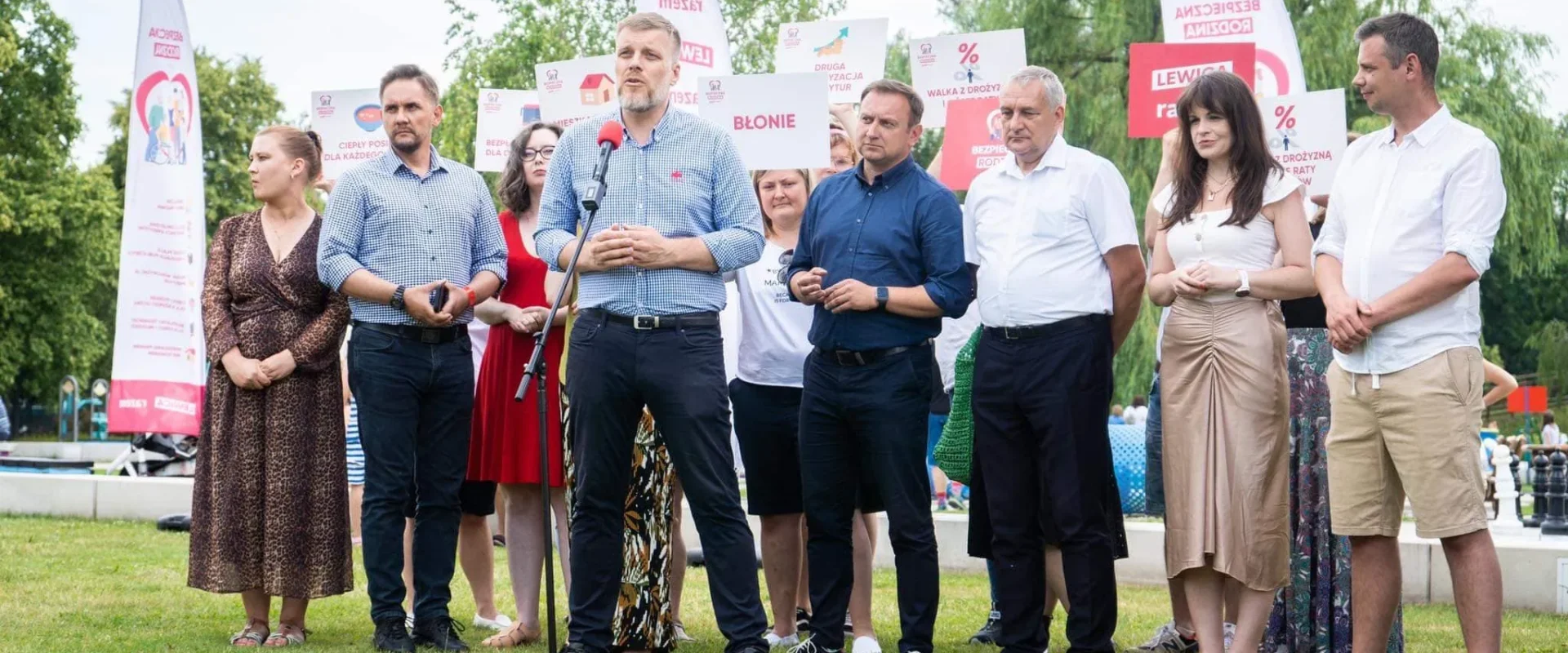 Na zdjęciu widoczna jest grupa osób stojących na zielonej trawie, z jednym mężczyzną mówiącym do mikrofonu, podczas wydarzenia plenerowego z banerami i transparentami.