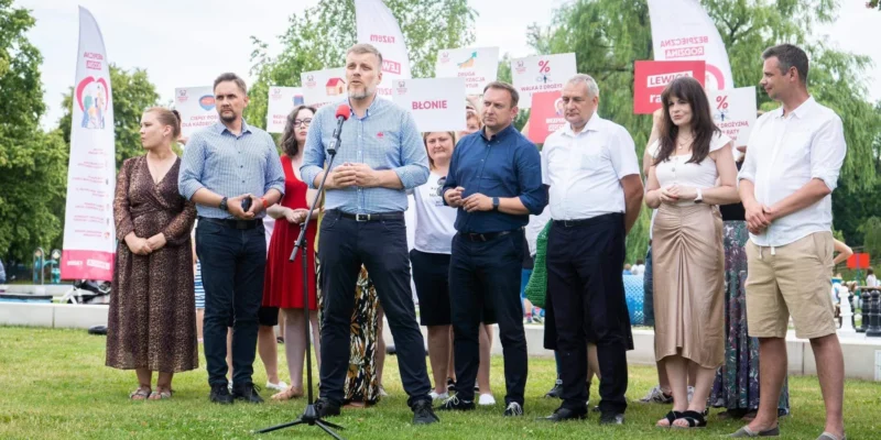 Na zdjęciu widoczna jest grupa osób stojących na zielonej trawie, z jednym mężczyzną mówiącym do mikrofonu, podczas wydarzenia plenerowego z banerami i transparentami.