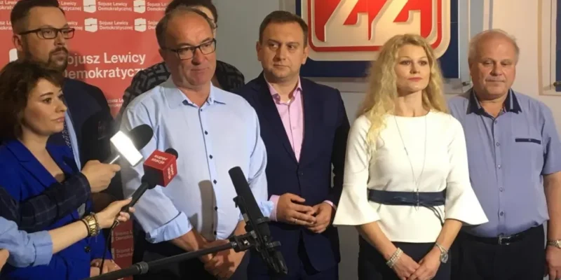 Kilka osób związanych z SLD Lewica Razem stoi przed mikrofonami i dużym logo partii podczas konferencji prasowej.