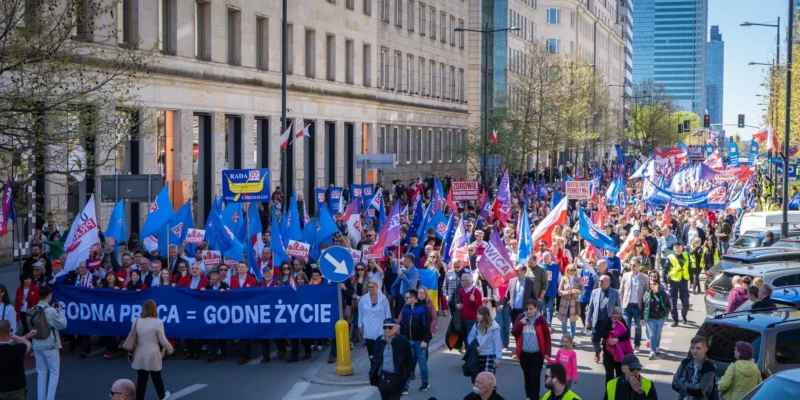 Tłumaczony marsz z transparentami i flagami, podczas którego uczestnicy wyrażają swoje postulaty dotyczące pracy i życia.