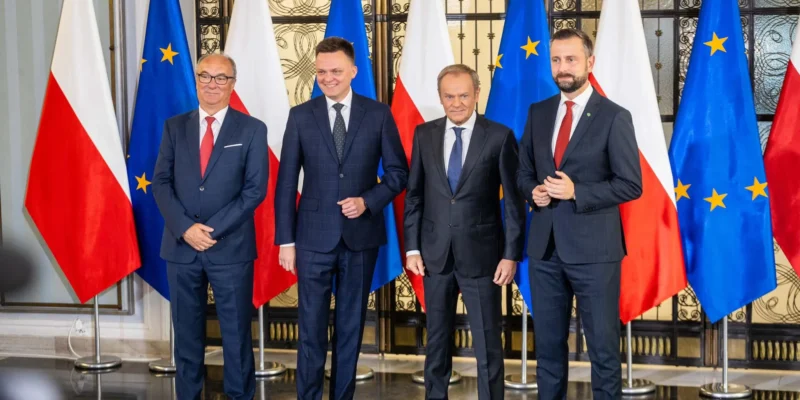 Czterech mężczyzn w garniturach stoi przed flagami Polski i Unii Europejskiej, prezentując wspólne oświadczenie opozycji.