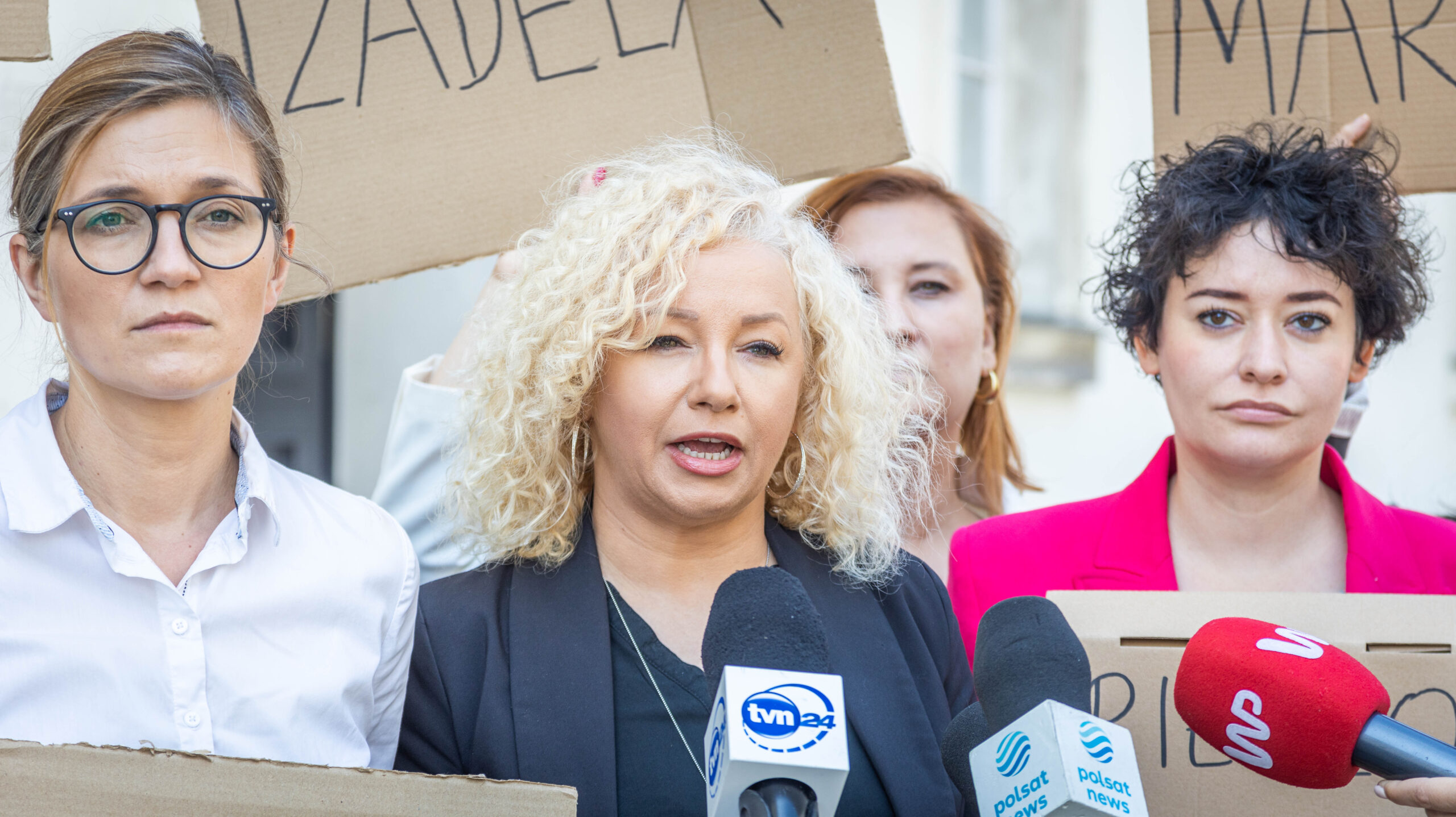 Grupa kobiet na proteście, jedna z nich przemawia do kamer mediów, stojąc przed kartonowymi transparentami.