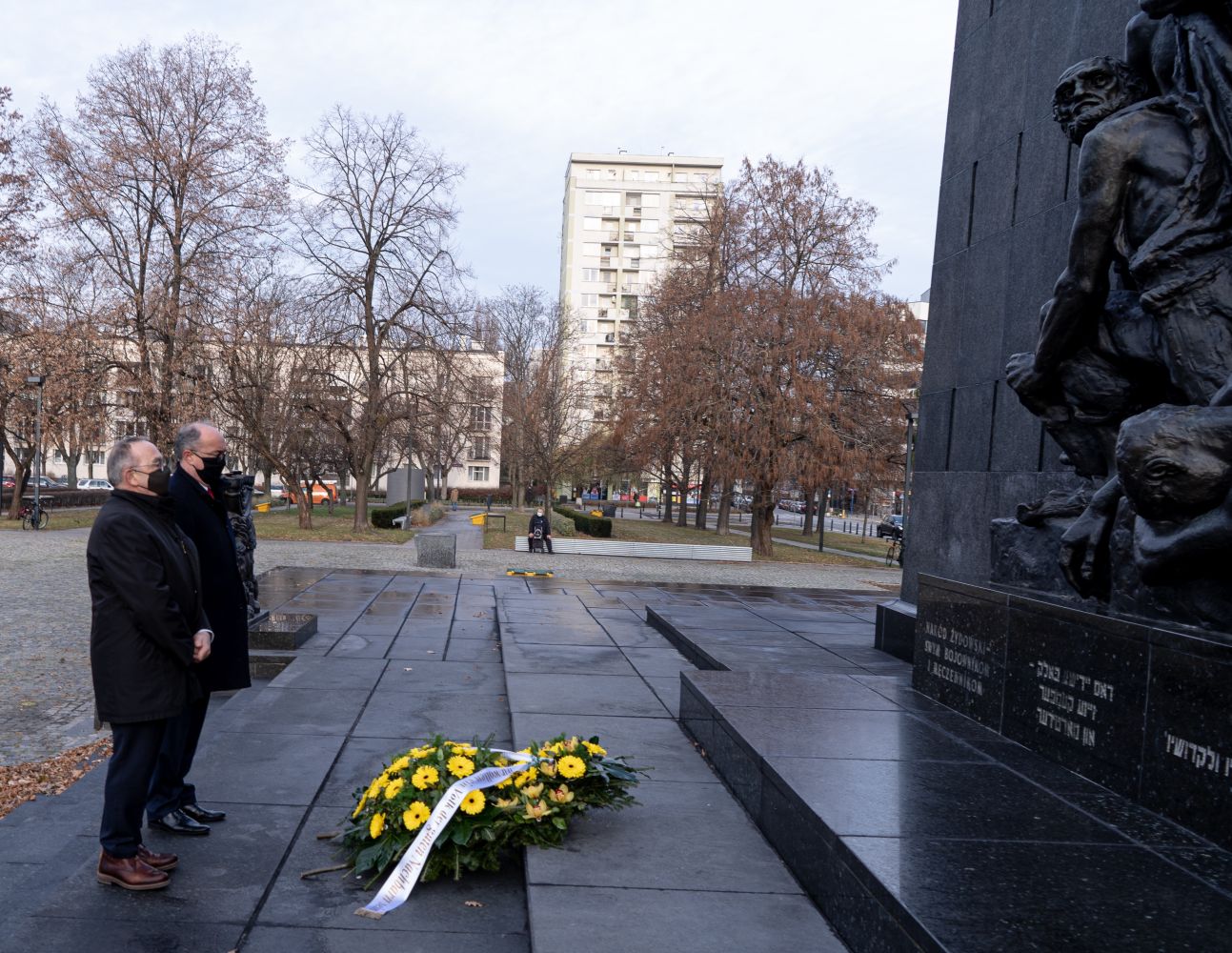 Dwóch mężczyzn w maseczkach stoi obok czarnego pomnika, obok którego leży wieniec z żółtymi kwiatami.
